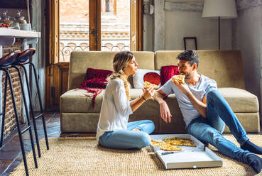 Young couple sitting on floor of living room and eating pizza from box - EHF00950