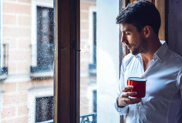 Junger Mann hält Tasse und schaut aus dem Fenster - EHF00974