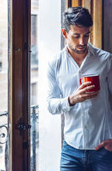 Nachdenklicher junger Mann mit Tasse am Fenster stehend - EHF00975