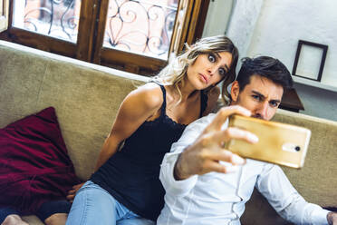 Young couple making faces while taking selfie on couch in living room at home - EHF00979