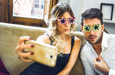 Verspieltes junges Paar, das eine Comedy-Brille trägt und ein Selfie auf der Couch im heimischen Wohnzimmer macht - EHF00980