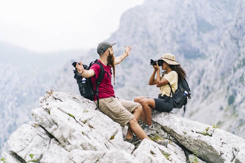 Junge Frau, die einen Mann fotografiert, während sie auf einem Berggipfel an der Ruta Del Cares sitzt, Asturien, Spanien - DGOF01416