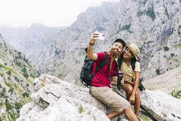 Lächelndes Paar, das ein Selfie macht, während es auf einem Felsen an der Ruta Del Cares sitzt, Asturien, Spanien - DGOF01418