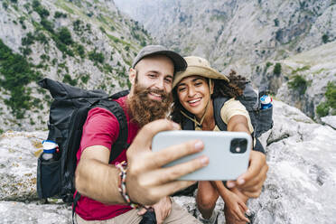 Couple smiling while taking selfie sitting on mountain peak at Ruta Del Cares, Asturias, Spain - DGOF01419