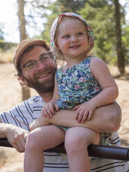 Vater hält seine Tochter auf dem Arm, während er in einem öffentlichen Park auf einer Stange sitzt - LAF02471