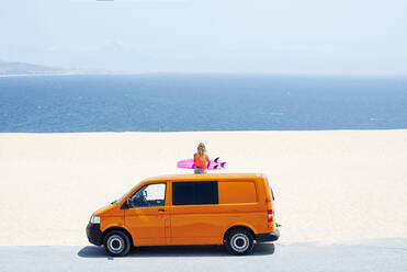 Junge Frau sitzt auf dem Dach eines Lieferwagens und schaut auf das Meer während der Sommerferien - JCMF01316