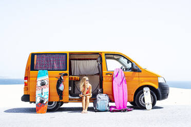 Woman sitting with hand on chin against van doorway at beach during sunny day - JCMF01329