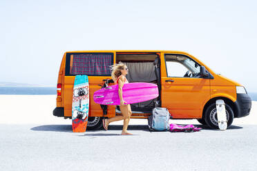 Frau mit wehenden Haaren läuft am Strand mit Surfbrett gegen Van an einem sonnigen Tag - JCMF01330