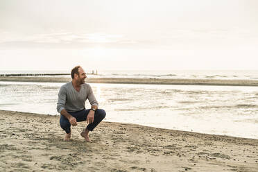 Älterer Mann lachend, während er am Strand gegen den Himmel bei Sonnenuntergang hockt - UUF21193