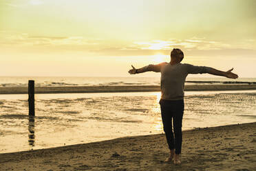 Carefree man with arms outstretched standing at beach against sky during sunset - UUF21194