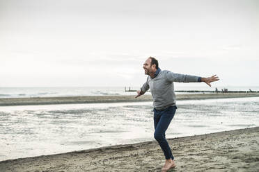 Unbekümmerter reifer Mann mit ausgestreckten Armen tanzt am Strand bei Sonnenuntergang - UUF21198