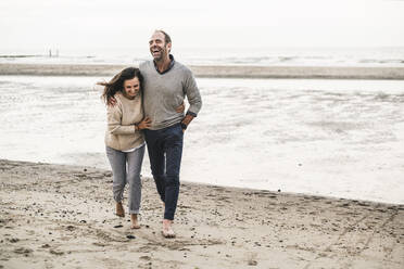 Cheerful couple walking at beach against sky during sunset - UUF21204