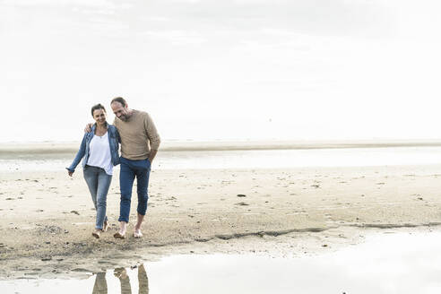 Smiling mature couple walking at beach against sky during sunset - UUF21212