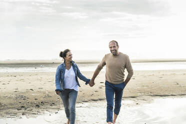 Mature couple holding hands while walking against sea during sunset - UUF21215