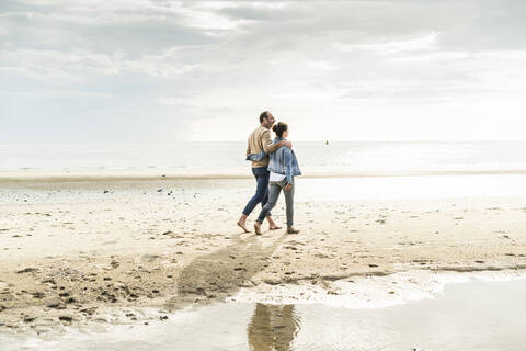 Paar mit Armen um zu Fuß am Strand gegen bewölkten Himmel bei Sonnenuntergang, lizenzfreies Stockfoto