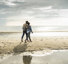 Paar mit Armen um Spaziergang am Strand gegen bewölkten Himmel während des Wochenendes - UUF21219