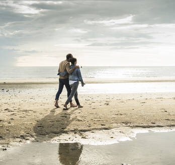 Couple with arms around walking at beach against cloudy sky during weekend - UUF21219