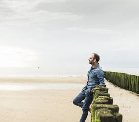 Thoughtful man standing by wooden posts at beach against sky during sunset - UUF21222