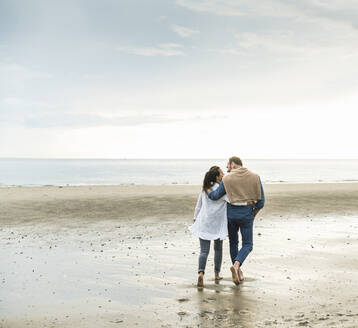 Mature couple with arms around enjoying weekend at beach against cloudy sky - UUF21229
