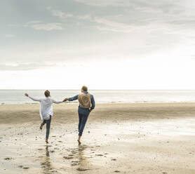Cheerful mature couple holding hands while jumping at beach against cloudy sky - UUF21230