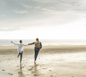 Fröhliches reifes Paar hält sich an den Händen, während es am Strand gegen den bewölkten Himmel springt - UUF21230