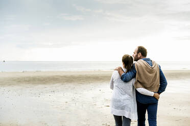 Couple with arms around looking at sea against cloudy sky during weekend - UUF21231