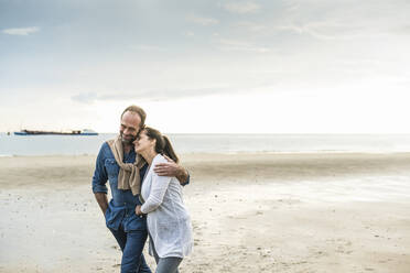 Mature man embracing woman while walking at beach against cloudy sky - UUF21233