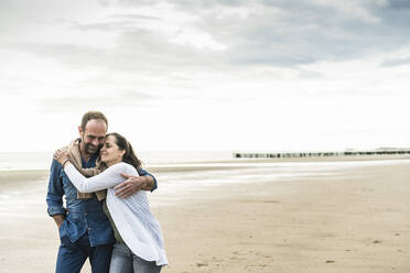 Mature woman embracing man while standing at beach against sky during sunset - UUF21234