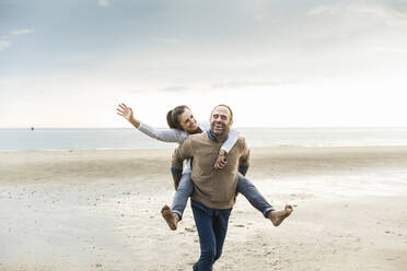 Happy mature man piggybacking woman while walking at beach against cloudy sky - UUF21235