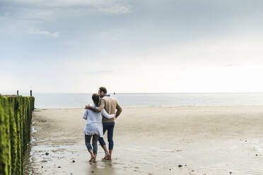 Loving couple with arms around walking at beach against cloudy sky - UUF21237