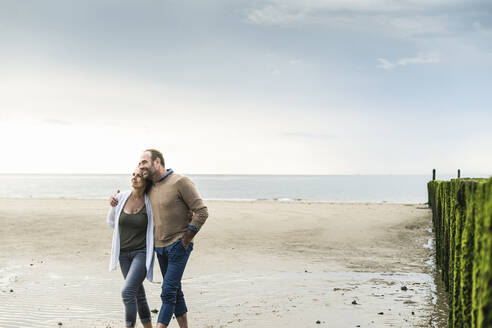 Loving mature couple walking at beach against sea during sunset - UUF21238