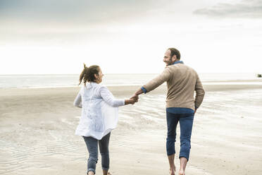 Unbekümmertes Paar hält sich an den Händen, während es bei Sonnenuntergang am Strand läuft - UUF21239