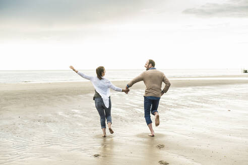 Fröhliches Paar hält sich beim Laufen am Strand während des Sonnenuntergangs an den Händen - UUF21240