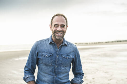 Fröhlicher Mann lachend am Strand stehend gegen den Himmel bei Sonnenuntergang - UUF21249