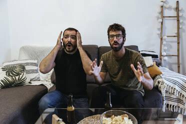 Stressed male friends watching sports while sitting on sofa at home - XLGF00487