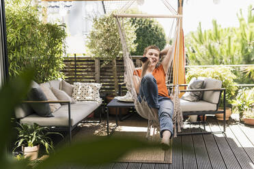 Cheerful mid adult woman talking over smart phone while swinging in porch seen through indoors - UUF21253