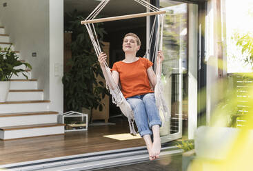 Mittlere erwachsene Frau mit geschlossenen Augen, die sich auf einer Schaukel vor einem Haus auf einer Veranda entspannt - UUF21259