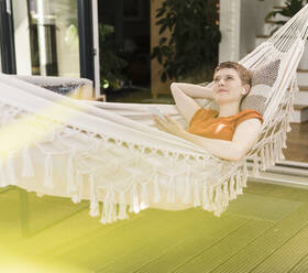 Thoughtful woman listening music through wireless headphones while lying on hammock in porch - UUF21264