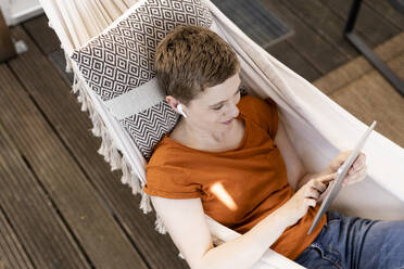 Mid adult woman with short hair using digital tablet while lying on hammock on porch - UUF21269