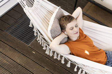 Woman with eyes closed listening music through wireless headphones while lying on hammock in porch - UUF21271