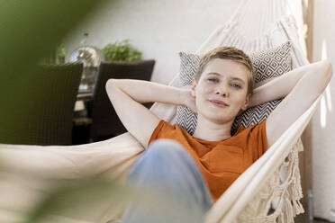 Smiling beautiful woman with hands behind head resting on hammock in porch - UUF21274