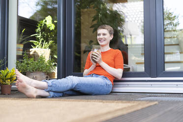 Smiling woman with short hair holding drink while sitting against house door in yard - UUF21289