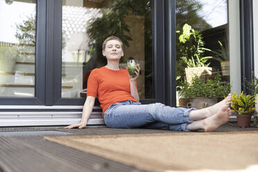 Frau mit geschlossenen Augen, die ein Getränk in der Hand hält, während sie auf der Veranda vor der Haustür sitzt - UUF21290