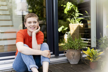 Smiling beautiful woman with hand on chin sitting against house in yard - UUF21293