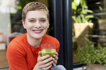 Close-up of smiling woman with short hair holding drinking glass while sitting against house - UUF21295