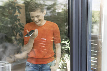 Lächelnde Frau, die über ein Mobiltelefon spricht, während sie zu Hause am Fenster steht, gesehen durch Glas - UUF21304