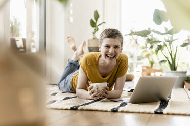 Cheerful woman holding coffee cup while using laptop on carpet at home - UUF21307