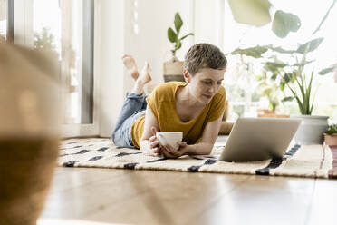 Mid adult woman holding coffee cup using laptop while lying on carpet at home - UUF21308