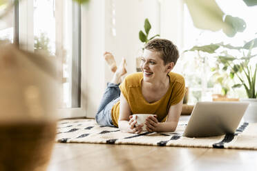 Cheerful woman looking away while lying with coffee cup and laptop on carpet at home - UUF21309