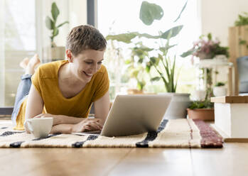 Lächelnde erwachsene Frau mit kurzen Haaren, die einen Laptop benutzt, während sie sich auf einem Teppich zu Hause entspannt - UUF21311